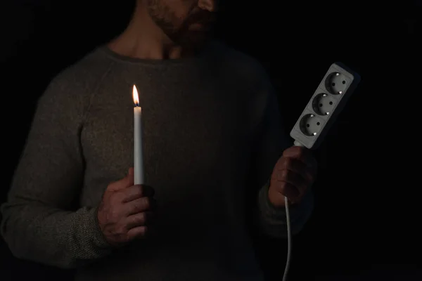 Vista parcial del hombre sosteniendo extensor de enchufe y vela encendida durante corte de energía aislado en negro - foto de stock