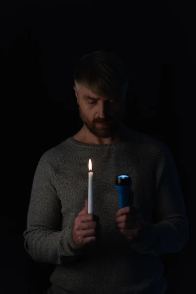 Человек в темноте, вызванной отключением электричества держа светящийся фонарик и зажженную свечу изолированы на черном — стоковое фото