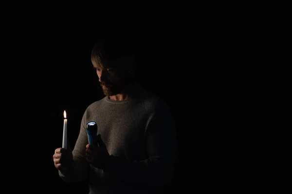 Man in darkness holding glowing flashlight and burning candle isolated on black — Stock Photo