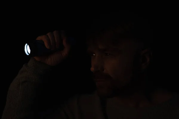 Man looking away in darkness while holding bright flashlight isolated on black — Stock Photo