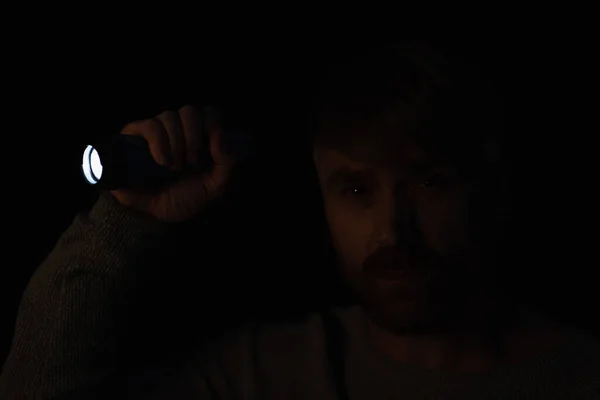 Mann im Dunkeln blickt in Kamera, während er helle Taschenlampe isoliert auf schwarz hält — Stockfoto