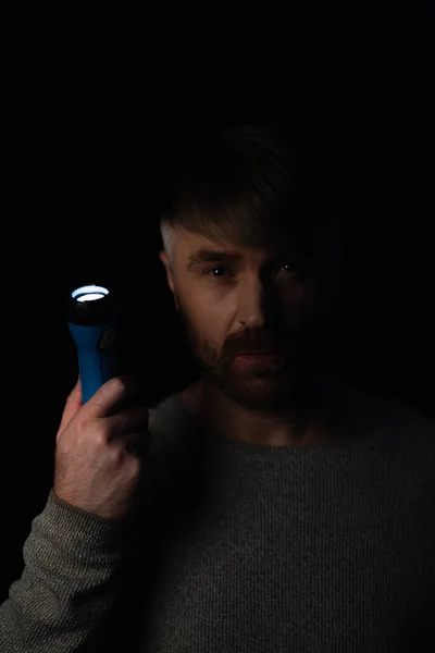 Hombre con linterna brillante mirando a la cámara durante el apagado de electricidad aislado en negro - foto de stock