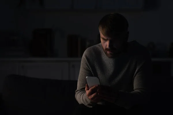 Hombre sentado en la cocina oscura y el uso de teléfono móvil durante el apagón de energía - foto de stock