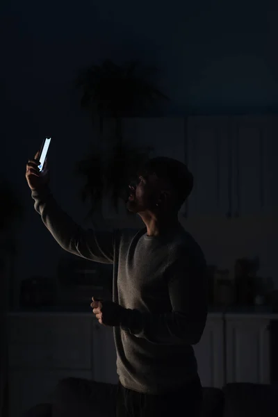 Man standing in dark kitchen with smartphone in raised hand and searching for connection during power blackout — Stock Photo