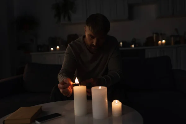 Homme allumant des bougies près du livre et du téléphone portable tout en étant assis dans la cuisine pendant une panne d'énergie — Photo de stock