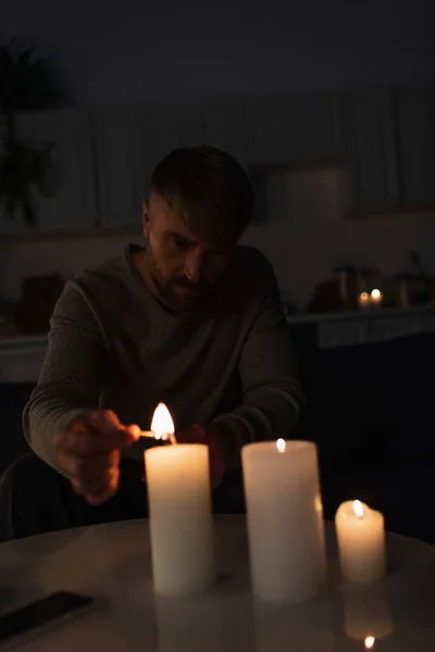 Man sitting in dark kitchen during power outage and lighting candles — Stock Photo