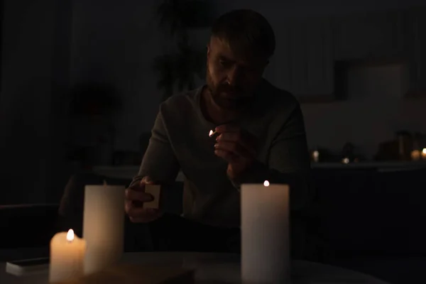 Человек держит зажженную спичку возле горящих свечей, сидя на кухне во время отключения электроэнергии — стоковое фото