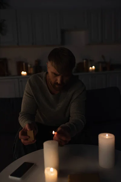 Hombre sentado en la cocina oscura durante el apagón de energía y encender velas - foto de stock