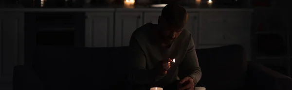 Чоловік з запаленим сірником сидить на кухні біля свічок під час відключення енергії, банер — стокове фото