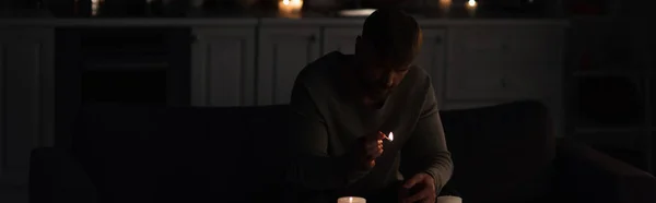 Man with lit match sitting in kitchen near candles during energy blackout, banner — Stock Photo