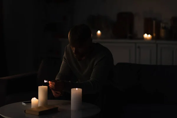 Man lighting candles in dark kitchen near mobile phone and book during energy blackout — Stock Photo