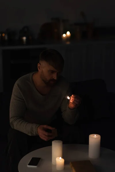 Человек смотрит на зажженный спичку, сидя в темной кухне возле горящих свечей во время отключения питания — стоковое фото