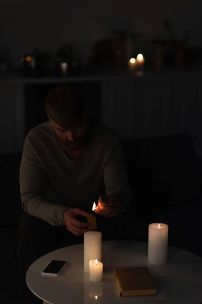Hombre sentado en la oscuridad y encendiendo velas cerca de libro y teléfono celular con pantalla en blanco - foto de stock