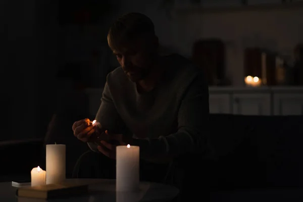 Uomo con fiammifero acceso seduto vicino alle candele e libro sul tavolo durante il blackout energetico — Foto stock