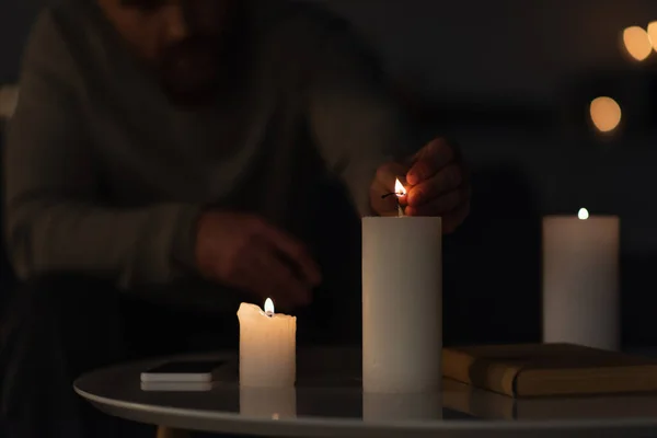 Partial view of man in darkness lighting candle near book and smartphone on table — Stock Photo