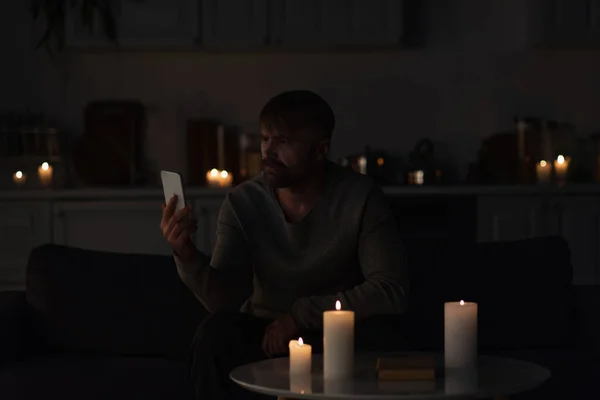 Homme regardant téléphone portable tout en étant assis dans la cuisine sombre près de bougies allumées — Photo de stock