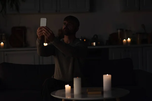 Человек сидит возле горящих свечей в темной кухне и ловит мобильное соединение на смартфоне — стоковое фото