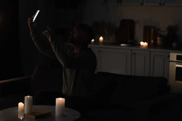 Mann sitzt in Dunkelheit neben brennenden Kerzen und fängt Handy-Verbindung mit Smartphone ein — Stockfoto