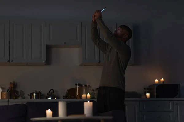 Чоловік тримає мобільний телефон у піднятих руках, ловлячи мобільний сигнал на темній кухні біля спалювальних свічок — стокове фото