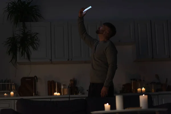 Чоловік стоїть на темній кухні з палаючими свічками і ловить мобільний зв'язок на мобільному телефоні — стокове фото