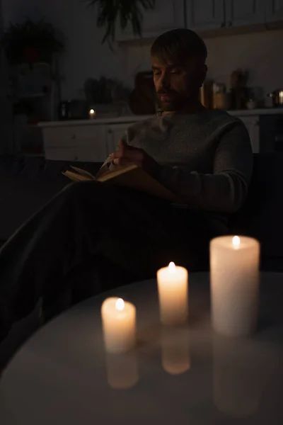 Человек читает книгу во время отключения электричества возле зажженных свечей в темной кухне — стоковое фото