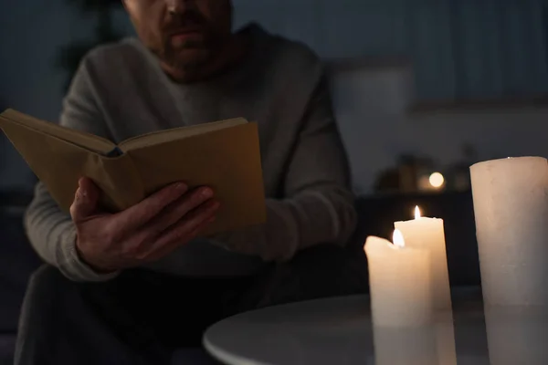 Cropped view of man reading book near candles burning in dark kitchen during power outage — Stock Photo