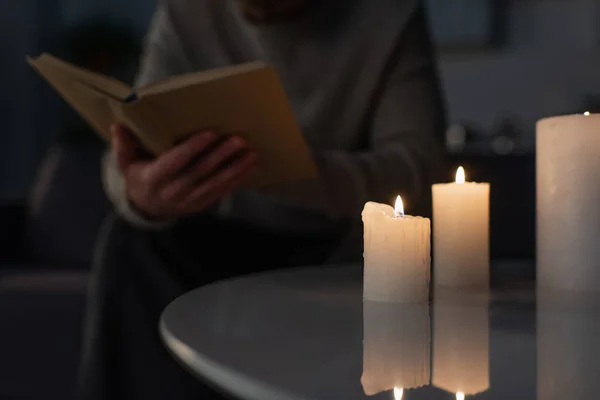 Partial view of blurred man reading book in darkness near burning candles on table — Stock Photo