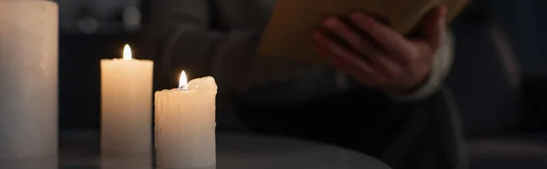 Selective focus of burning candles near cropped man reading book in darkness on blurred background, banner — Stock Photo
