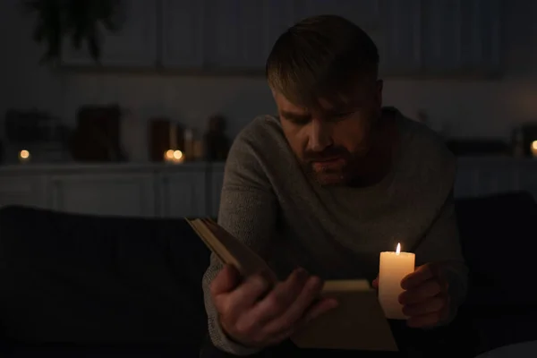 Man holding lit candle while reading book in dark kitchen during electricity outage — Stock Photo