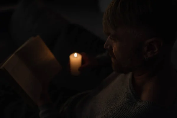 Человек держит горящую свечу во время чтения книги в темноте, вызванной отключением энергии — стоковое фото