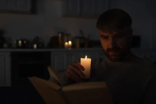Man holding lit candle while reading book in dark kitchen during electricity shutdown — Stock Photo