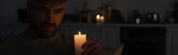 Man holding burning candle in dark kitchen during electricity outage, banner — Stock Photo