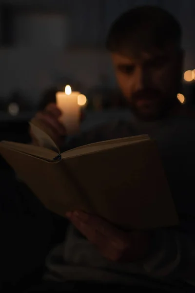 Foco seletivo do livro perto do homem segurando vela acesa durante a leitura durante o desligamento da eletricidade — Fotografia de Stock