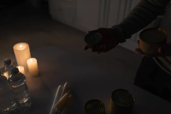 Vue partielle de l'homme tenant des canettes près de la table avec de l'eau embouteillée et des bougies dans la cuisine sombre — Photo de stock