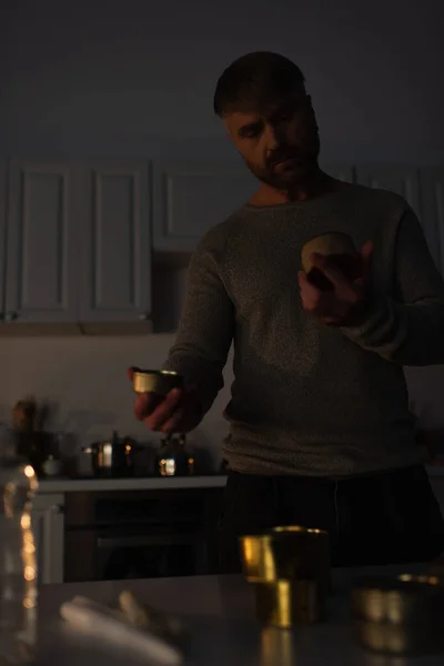 Uomo che tiene cibo in scatola in cucina scura mentre prepara riserva durante blackout energetico — Foto stock
