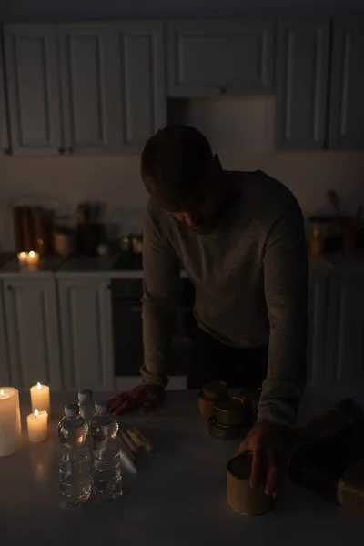 Man standing in dark kitchen near stock of canned food with bottled water and candles — Stock Photo