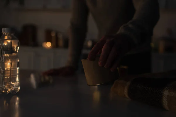 Vue recadrée de l'homme tenant de la nourriture en conserve près d'une couverture chaude et de l'eau embouteillée dans une cuisine sombre — Photo de stock