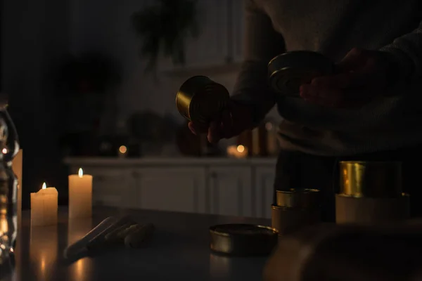 Partial view of man holding canned food near candles in kitchen during electricity shutdown — Stock Photo