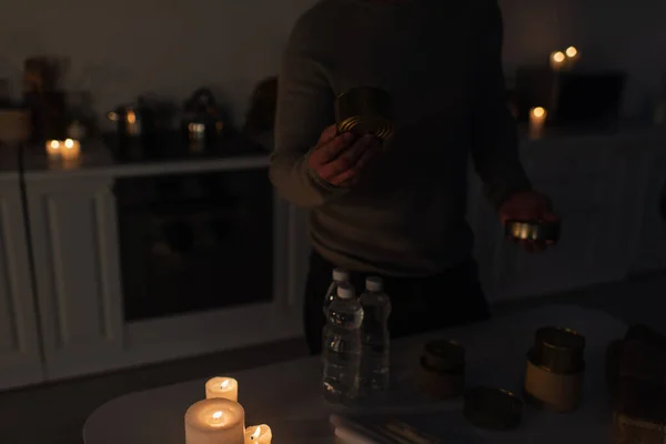 Обрезанный вид человека с консервами рядом с бутилированной водой и горящими свечами в темной кухне — стоковое фото