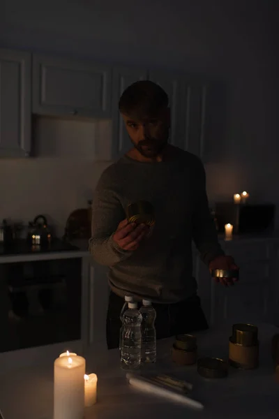 Человек смотрит в камеру на темной кухне, держа консервы возле стола с бутилированной водой и свечами — стоковое фото