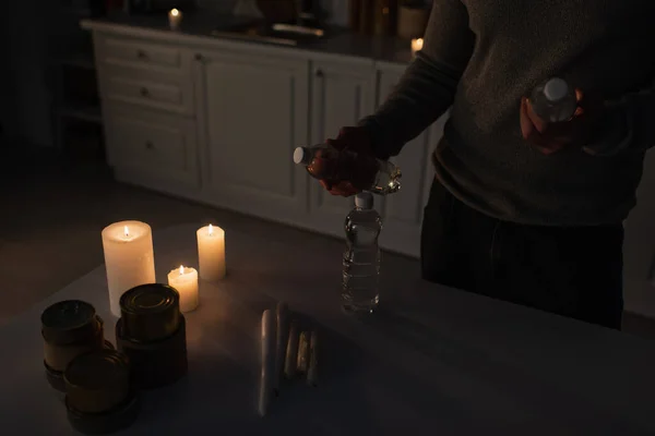 Cropped view of man with bottled water near table with canned food and candles in dark kitchen — Stock Photo