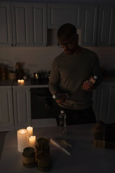 Man in eyeglasses holding bottled water near reserve of canned food and candles on table in dark kitchen — Stock Photo