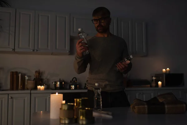 Людина в окулярах тримає пляшкову воду біля столу з консервованою їжею і теплим покриттям на темній кухні — стокове фото