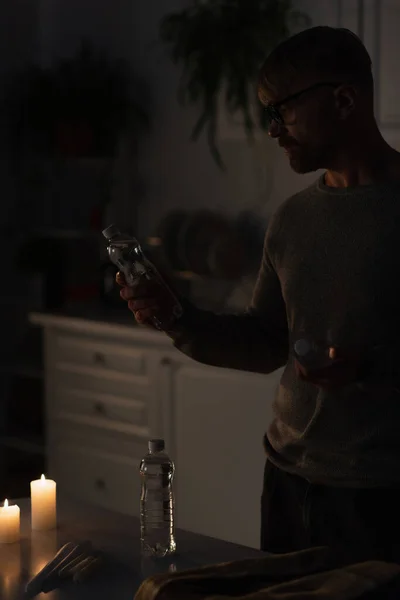 Man holding bottled water near burning candles in dark kitchen during electricity shutdown — Stock Photo