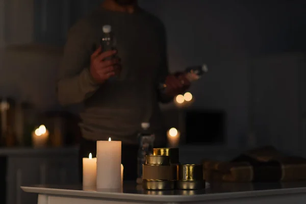 Cropped view of blurred man holding bottled water near table with stock of canned food near candles and warm blanket — Stock Photo