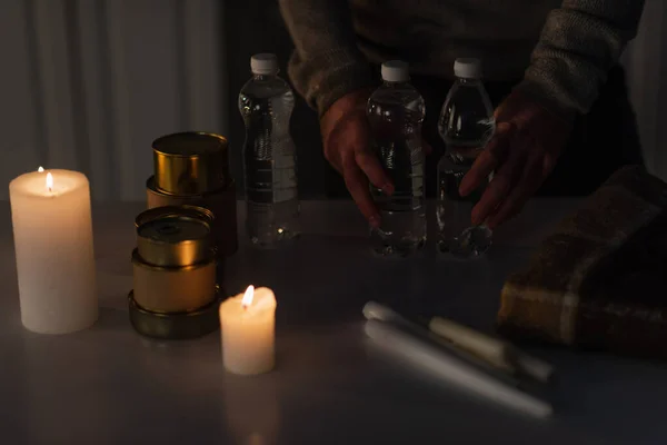 Vista ritagliata dell'uomo che tiene l'acqua in bottiglia vicino a cibo in scatola e candele durante il blackout energetico — Foto stock