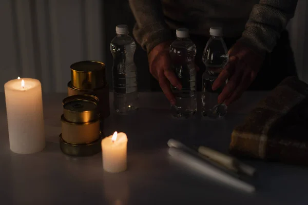 Cropped view of man holding bottled water near canned food and candles during energy blackout — Stock Photo