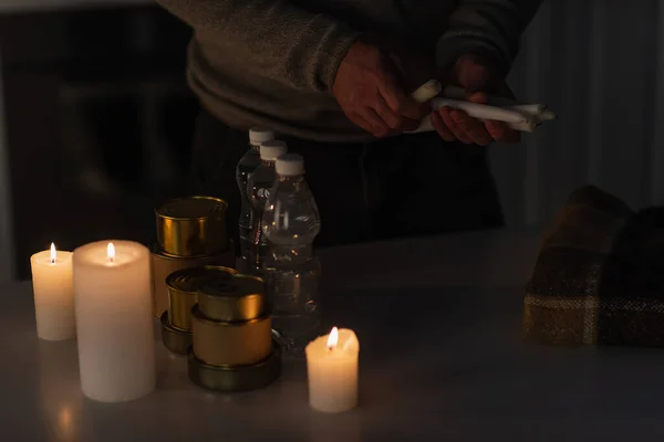 Partial view of man with candles near table with warm blanket and reserve of water and canned food — Stock Photo