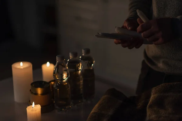 Частичный вид человека, держащего свечи возле бутилированной воды с консервами и теплым одеялом на столе — стоковое фото