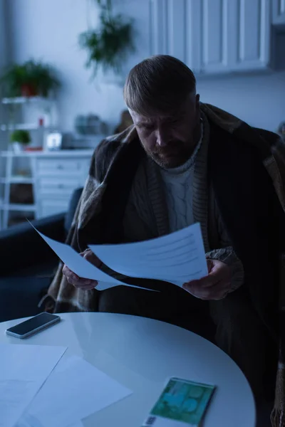 Чоловік сидить під теплим покриттям і дивиться на рахунки поблизу грошей і смартфона під час вимкнення живлення — стокове фото